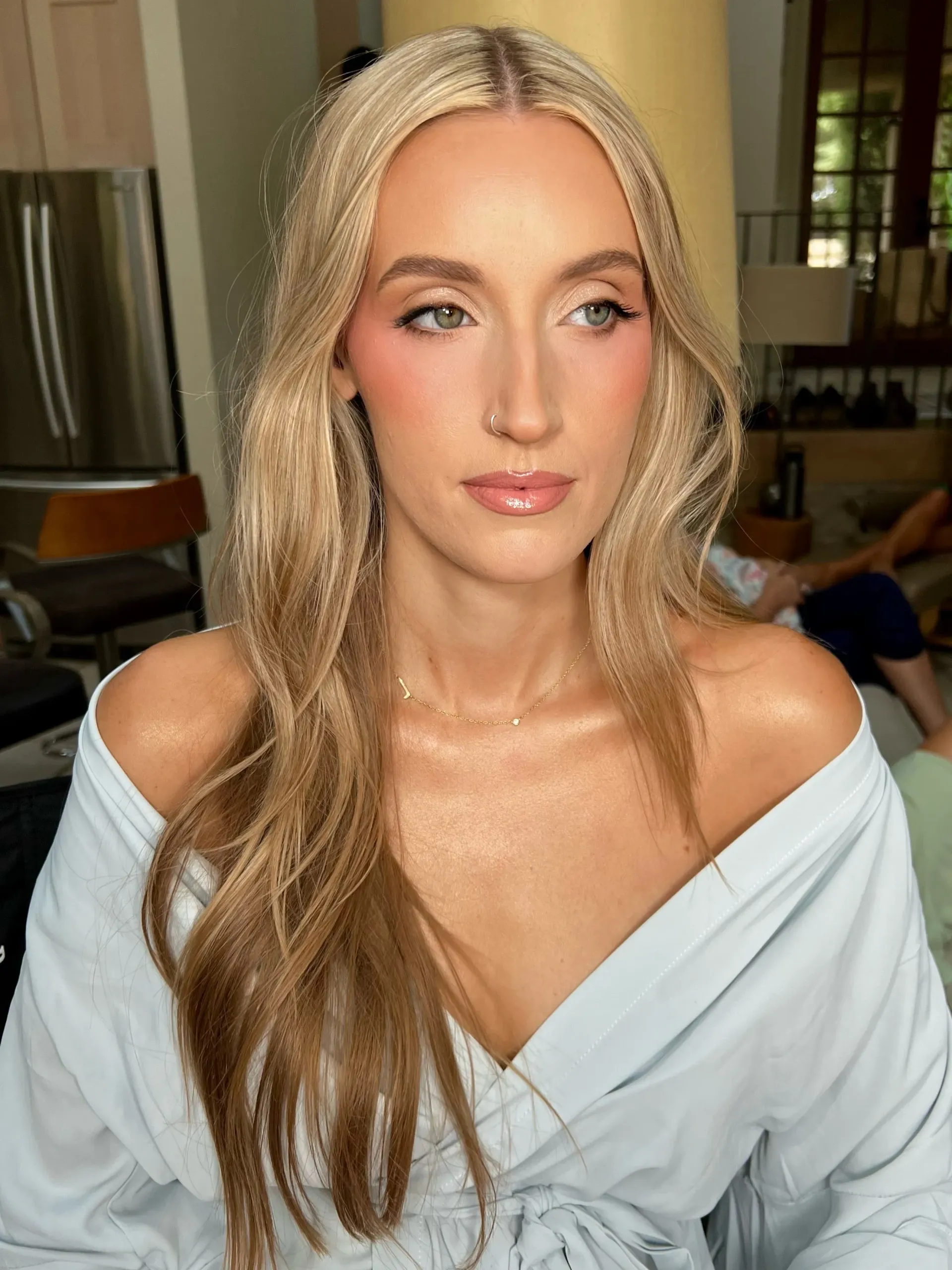 Woman with long blonde hair and light makeup, wearing a blue off-the-shoulder top. She is looking slightly to the side in a well-lit interior.
