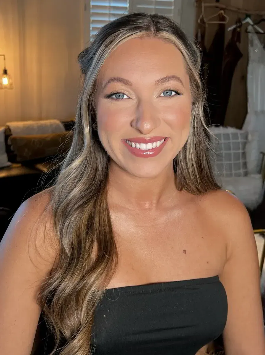 Woman with wavy blonde hair smiling, wearing a black strapless top indoors.