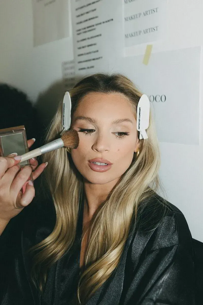 Woman with blonde hair, wearing a blue off-the-shoulder shirt, light makeup with pink blush. Eyes closed.