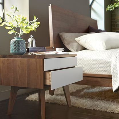 Nightstand with drawers, vase with greenery, and a cup, next to a bed with pillows.
