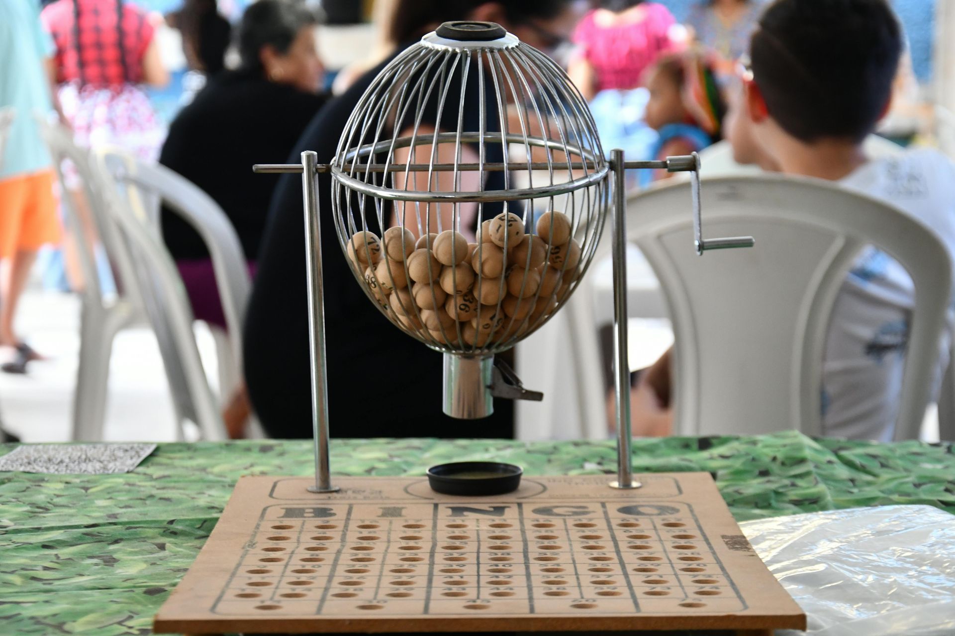 Máquina de loteria com bolas, sobre uma mesa. Pessoas ao fundo.