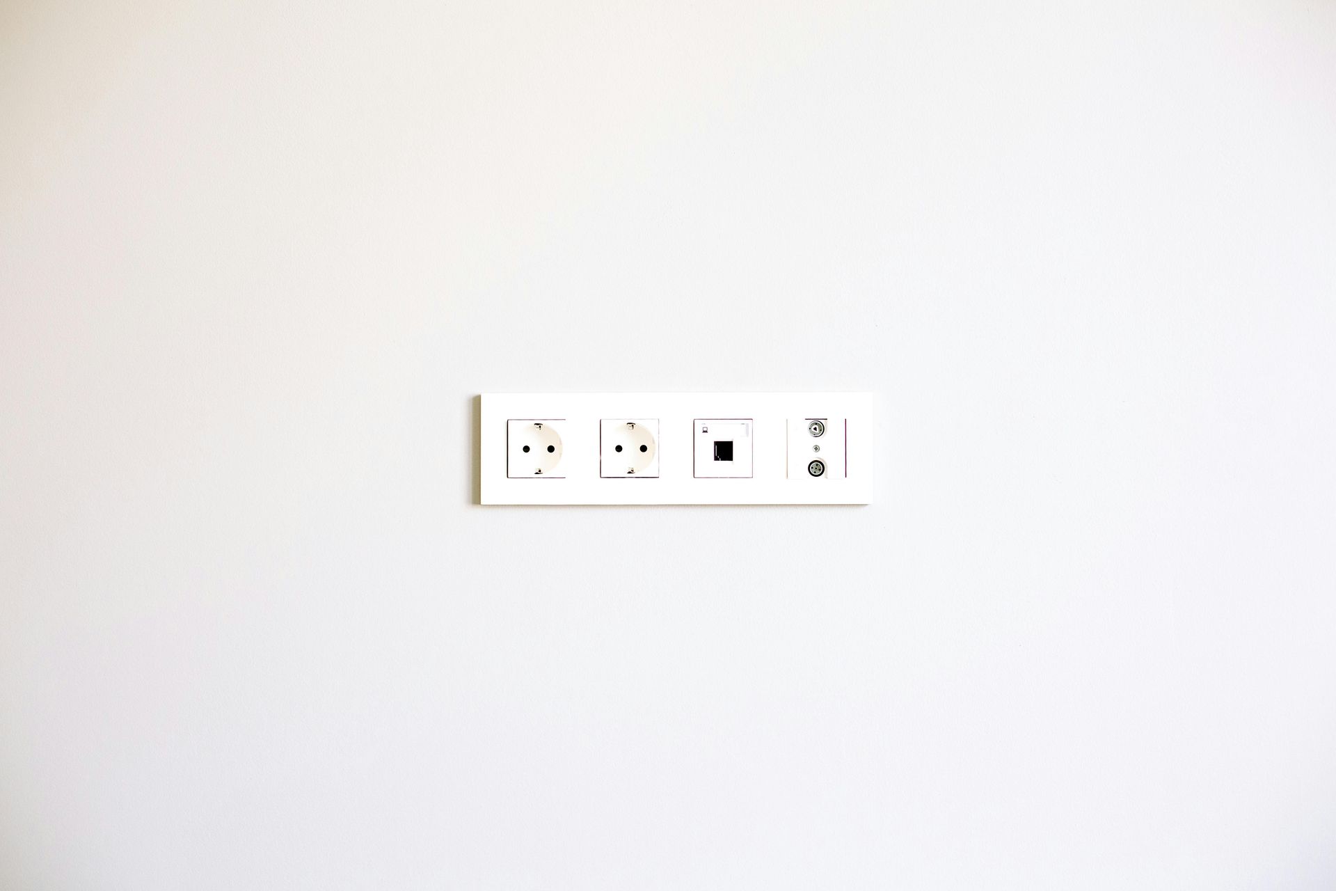 Parede branca com painel de tomadas retangular branco. Possui duas tomadas elétricas, uma entrada preta para telefone e uma porta branca para dados.
