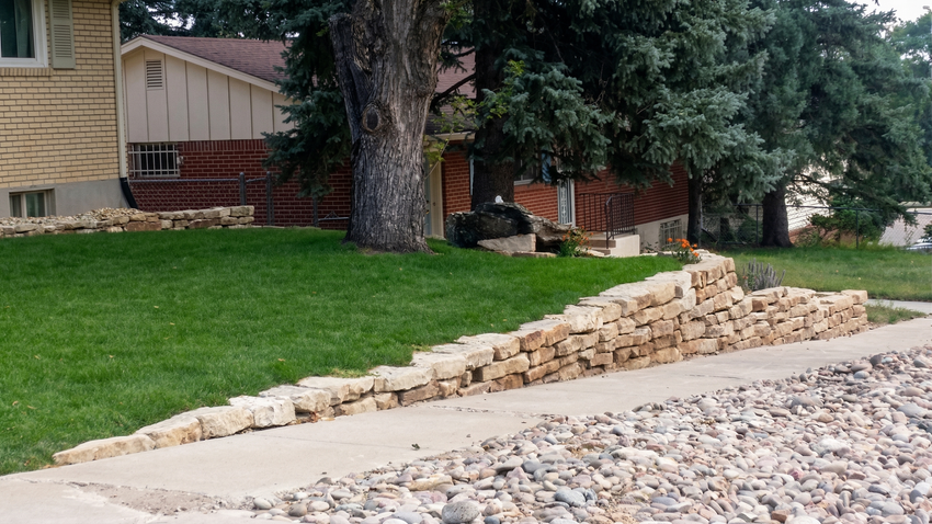 Stone retaining wall under construction beside a grassy yard and gravel driveway, with houses and trees in the background