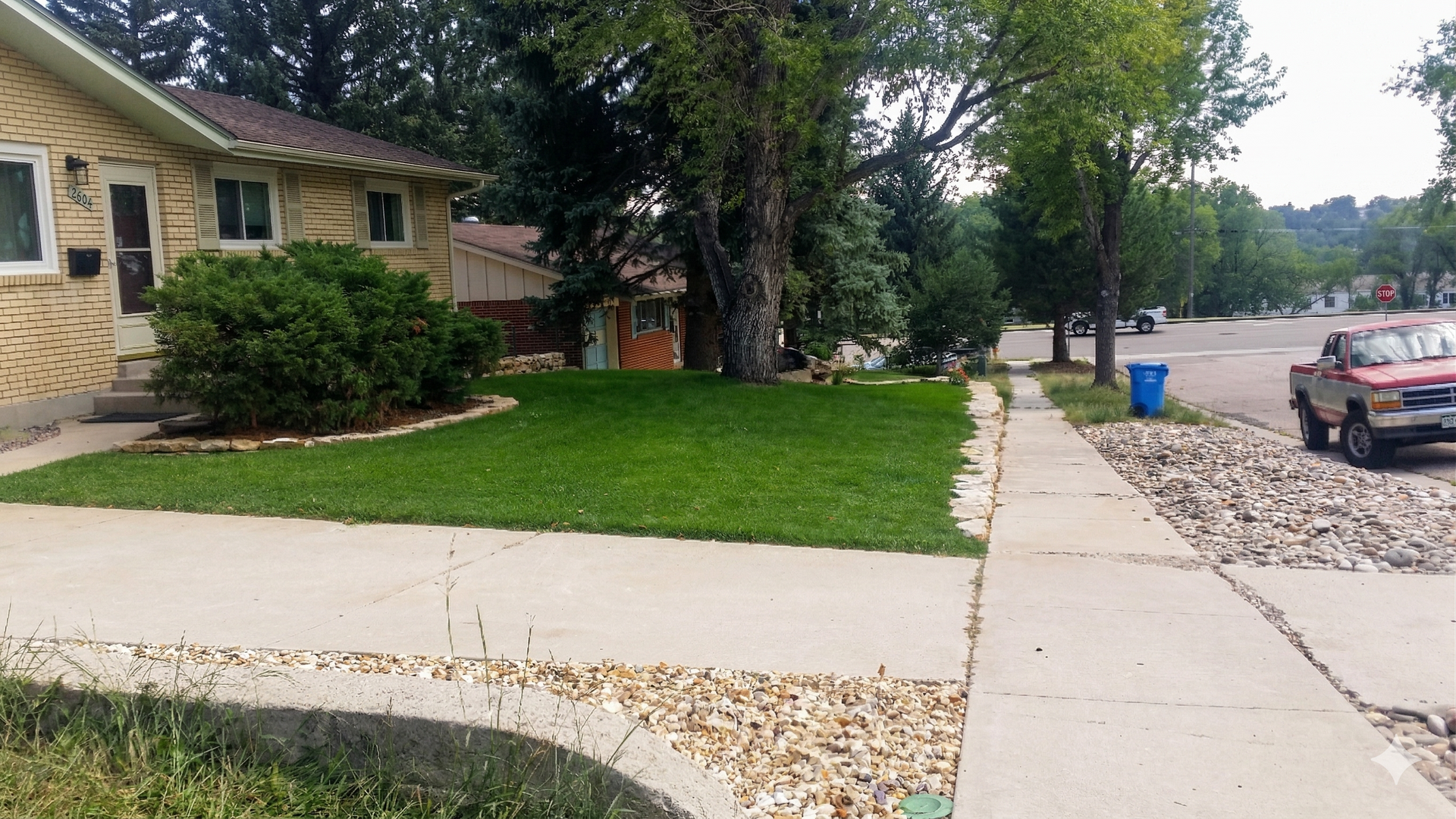 Suburban street with a stone house, green lawn, sidewalk, trees, and parked cars along the curb.