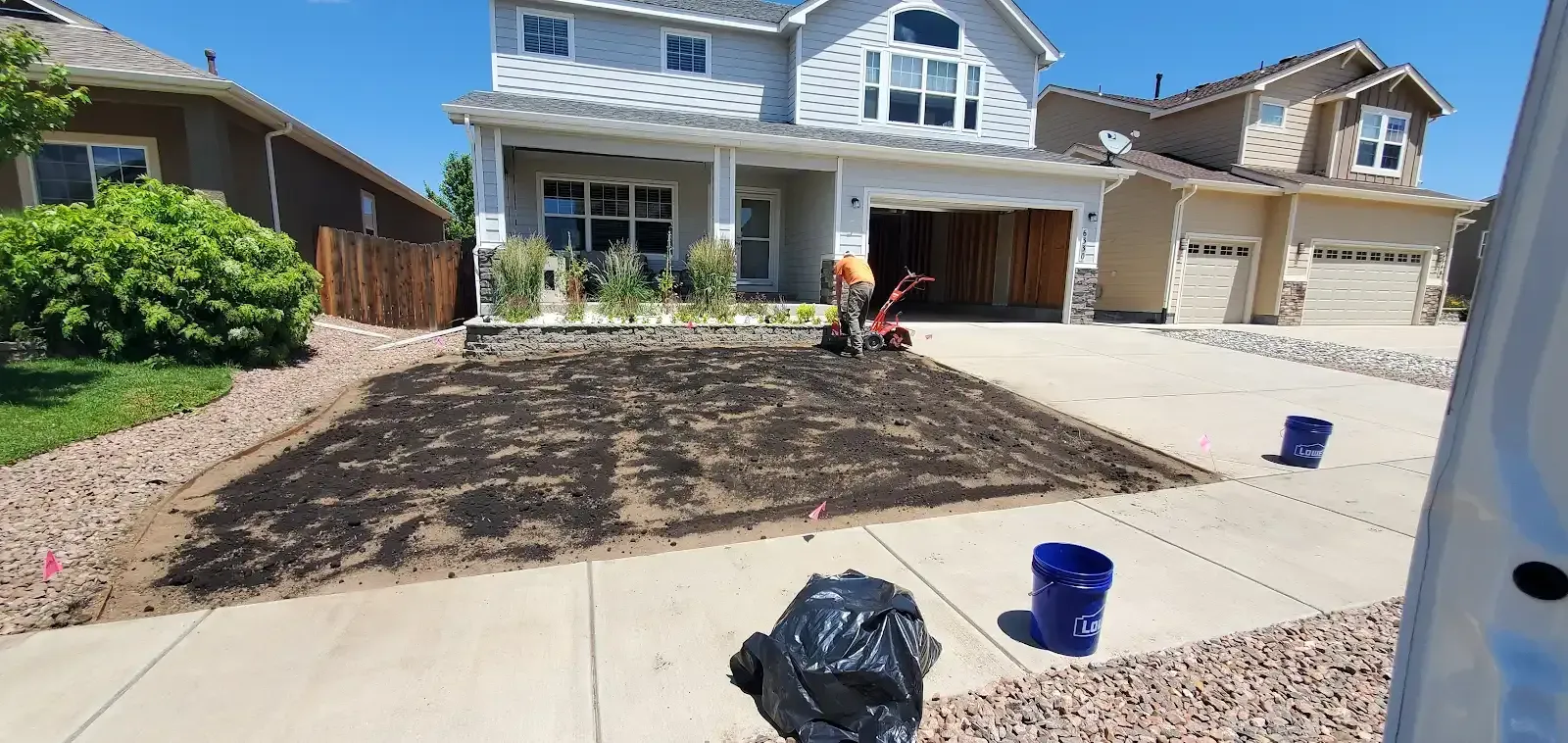 Workers preparing a front yard for landscaping at a suburban house, with bare soil and buckets in the driveway.