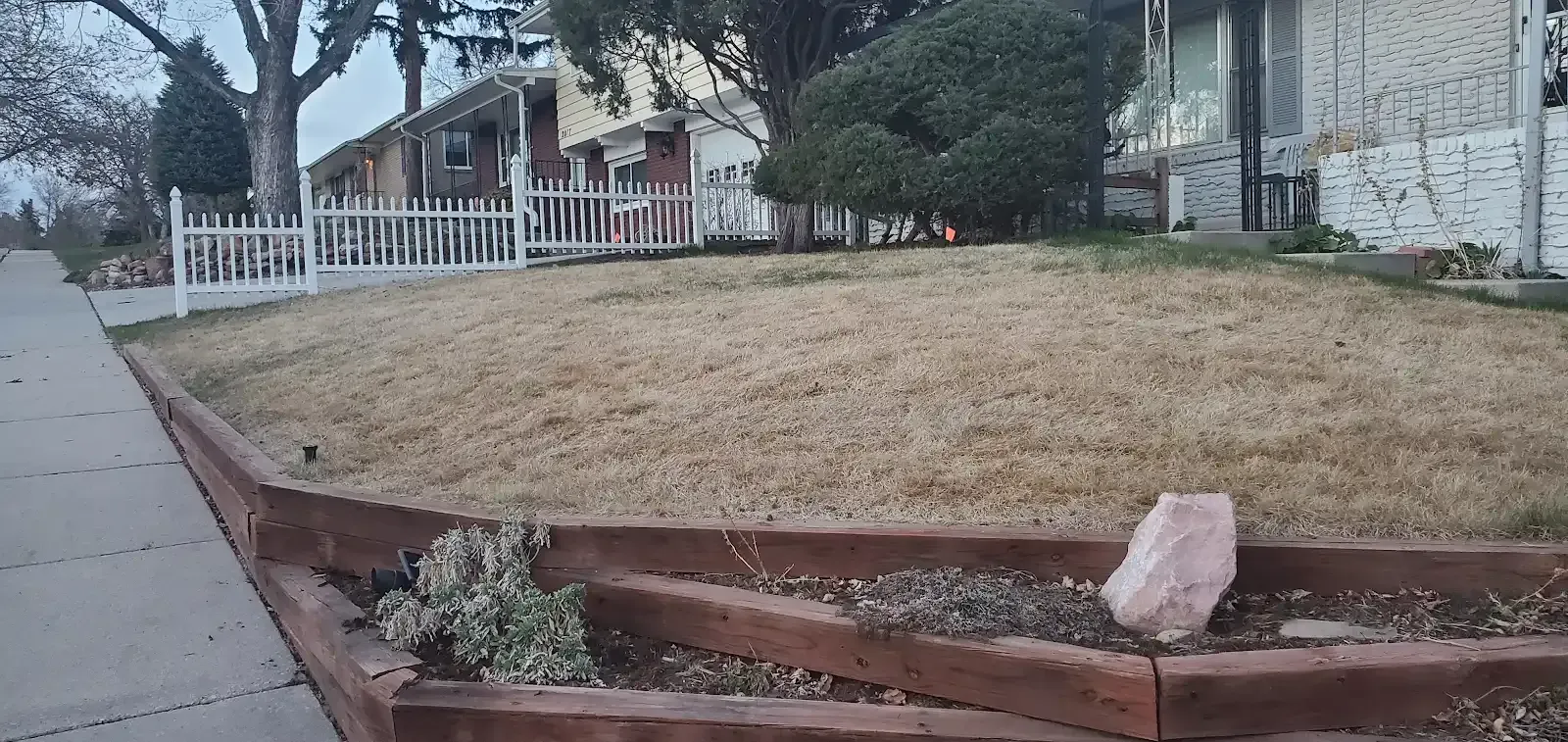 Flood-damaged front yard with eroded red curb and a muddy lawn beside a white fence and houses