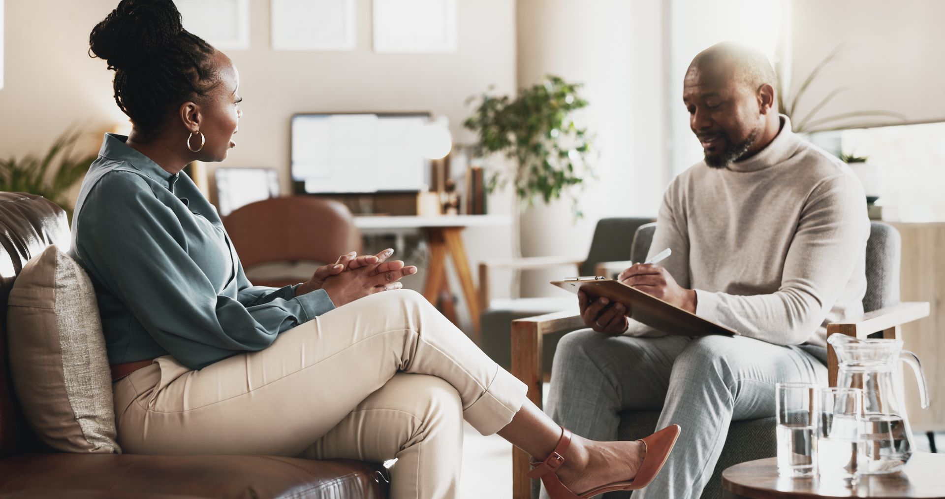 Two women sit in a modern office, holding hands and offering support while talking near a box of tissues.