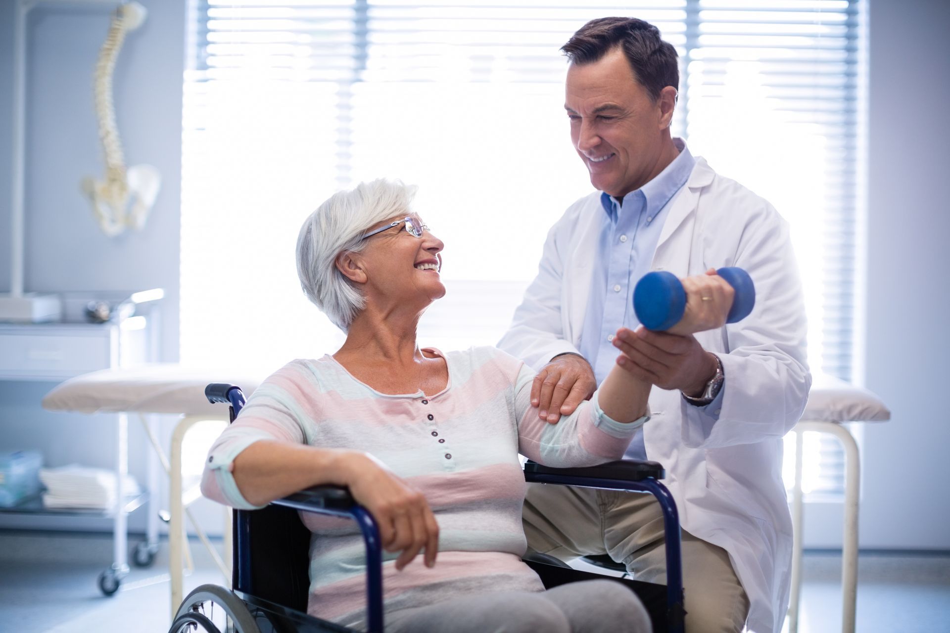 A healthcare professional assists a person in a wheelchair with an arm exercise using a small blue dumbbell in a clinic.