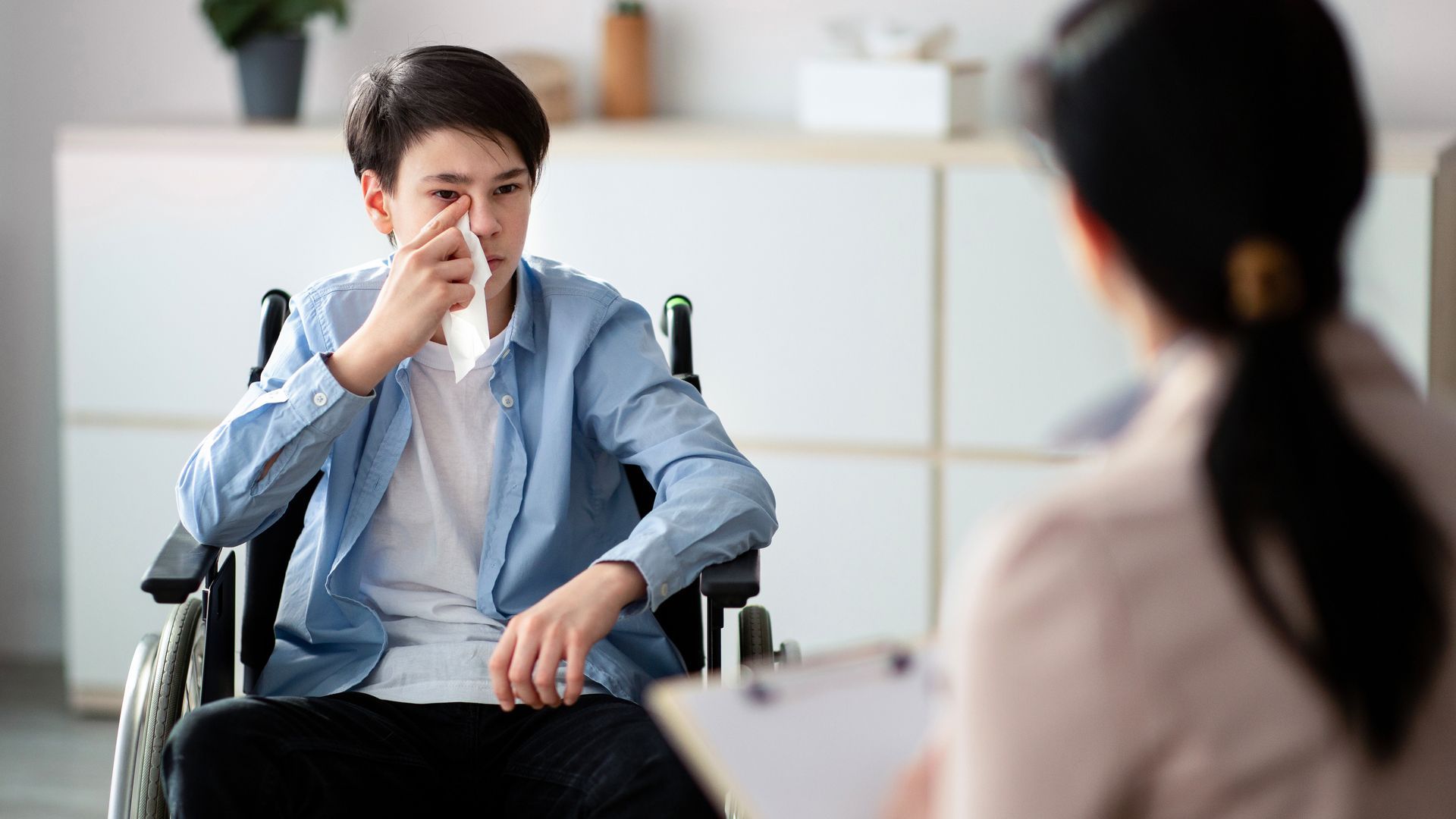 A person in a wheelchair uses a tissue to wipe their eye while speaking with a counselor in an office setting.
