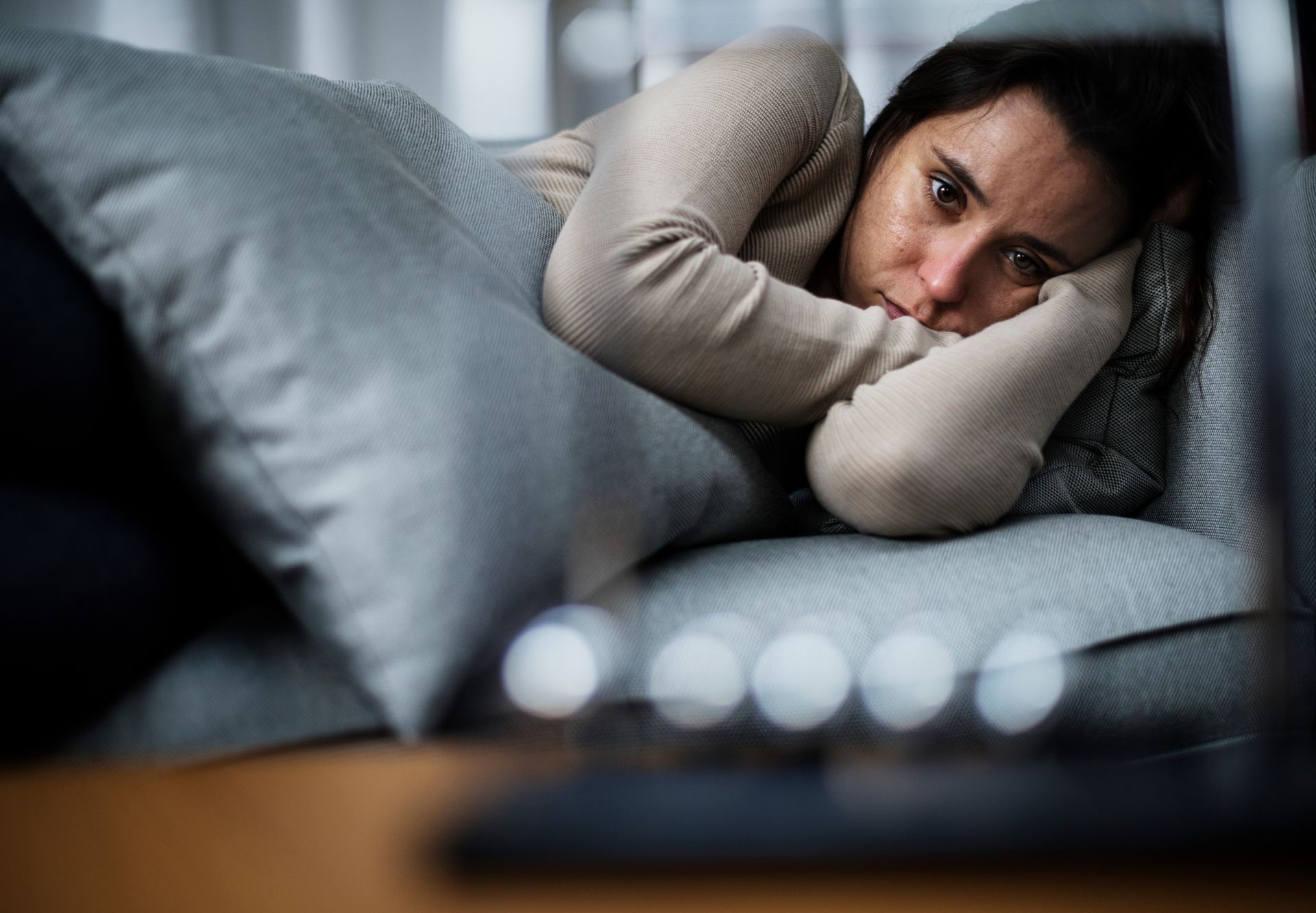 A person lying on a couch, resting their head on their arms, looking directly ahead with a somber expression.