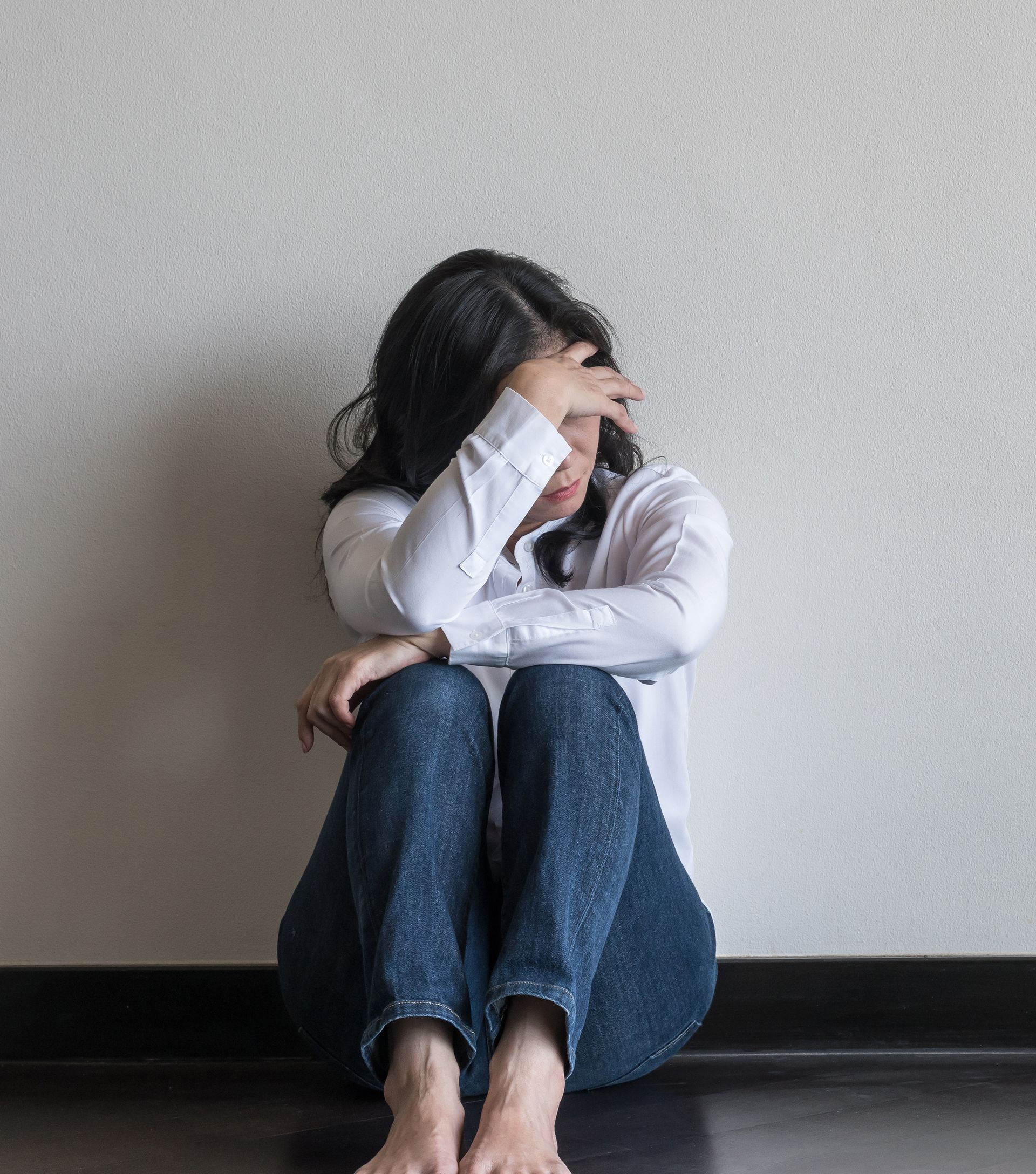 A person sits on the floor with their head in their hand, leaning against a neutral-colored wall.