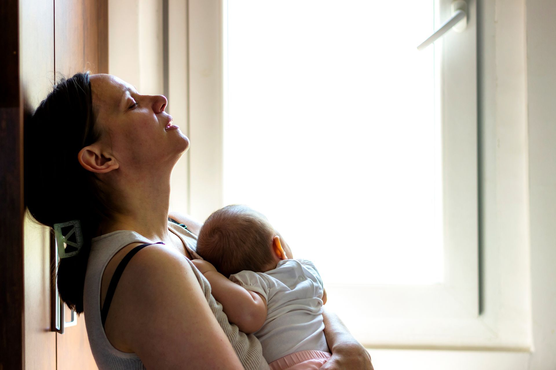 A person leans their head against a wall while holding an infant against their shoulder near a bright window.