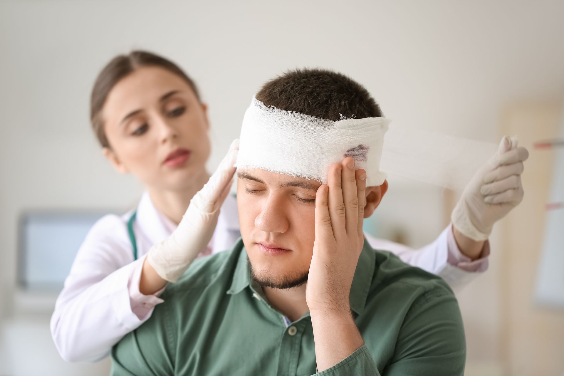 A medical professional in a white coat and gloves bandages a patient's head in a clinical setting.