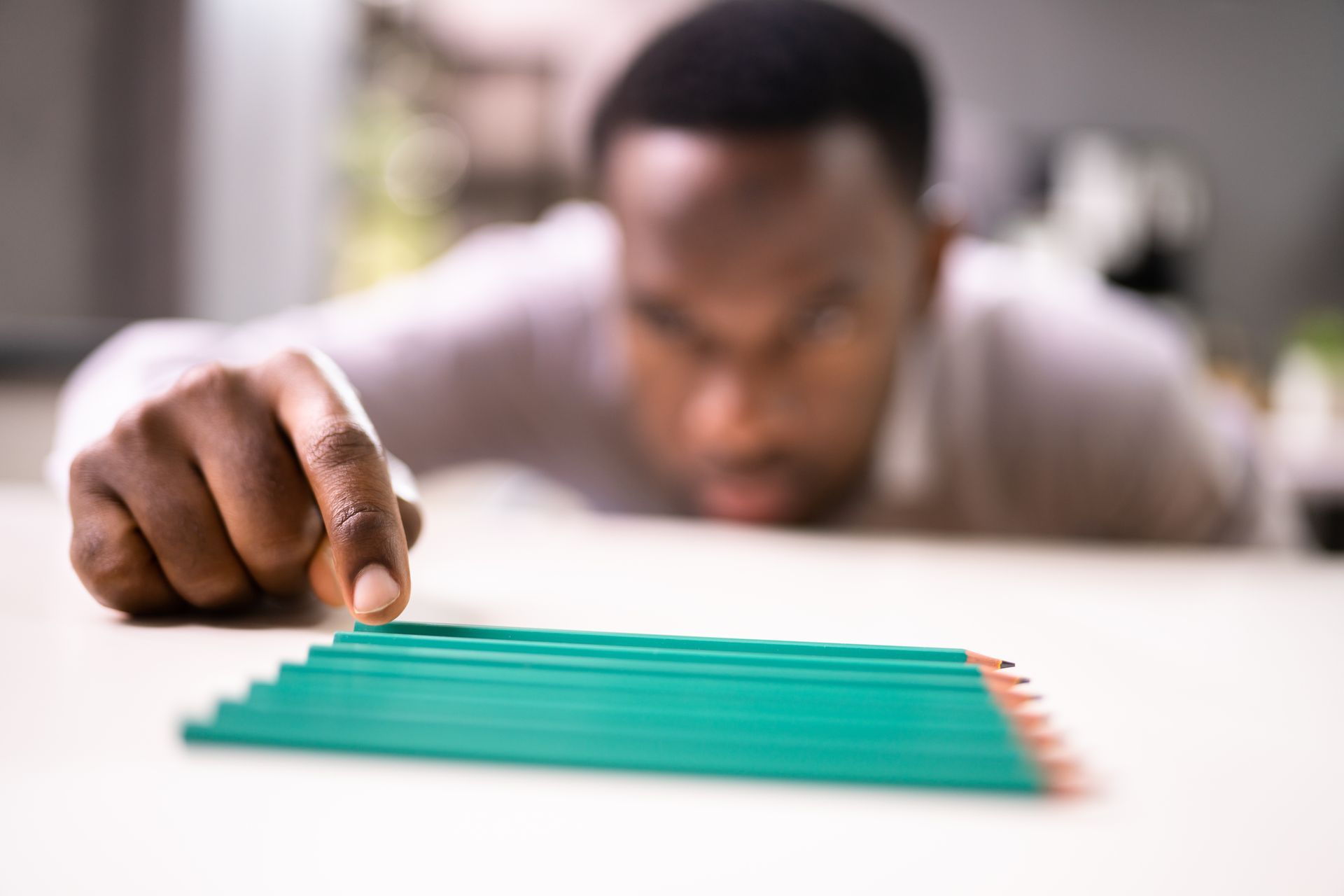 A person looks closely at a row of colorful pencils arranged on a reflective table, pointing to the red one.