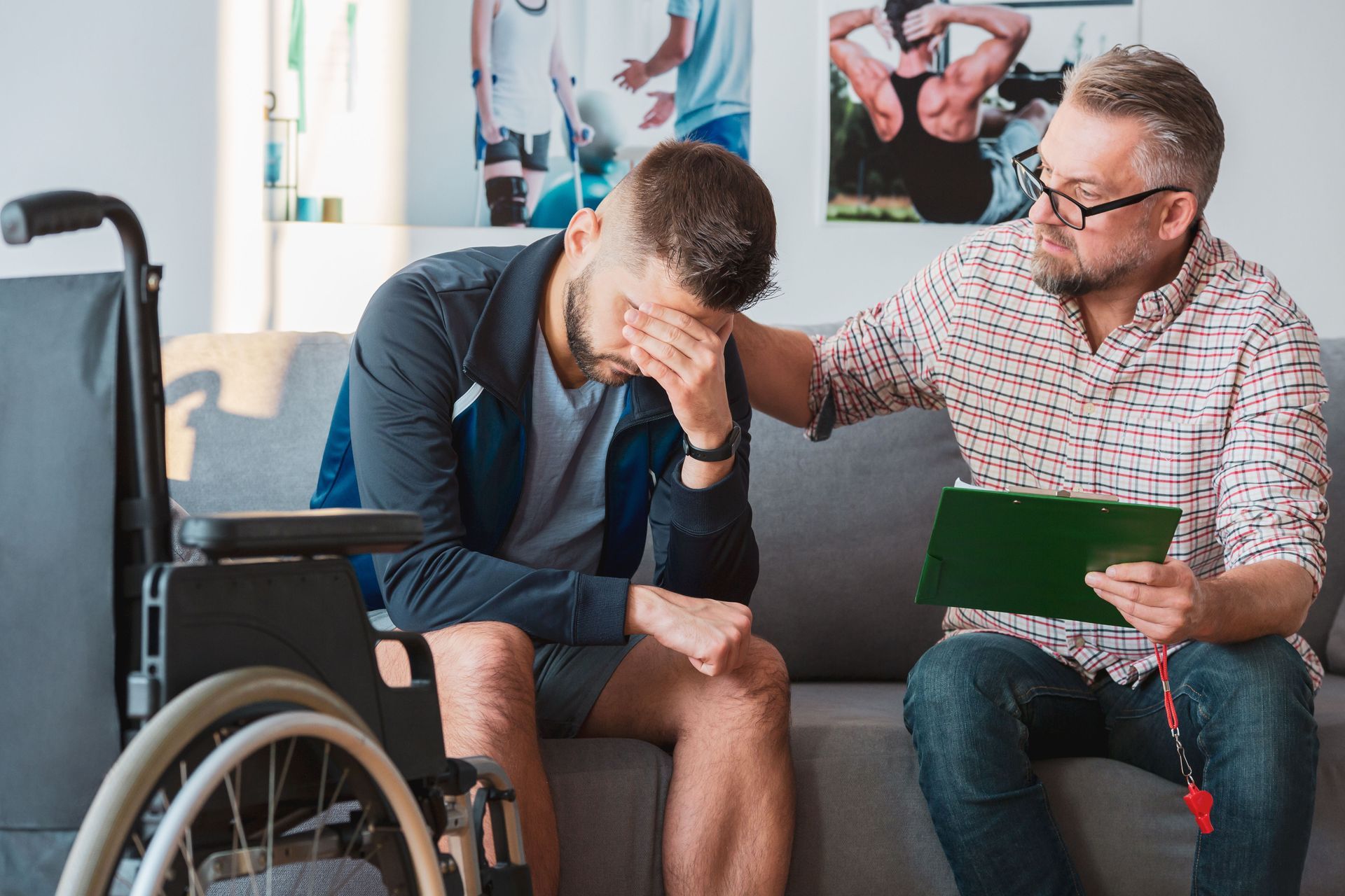 A man with his head in his hands sits on a couch next to a therapist who offers support, with a wheelchair nearby.