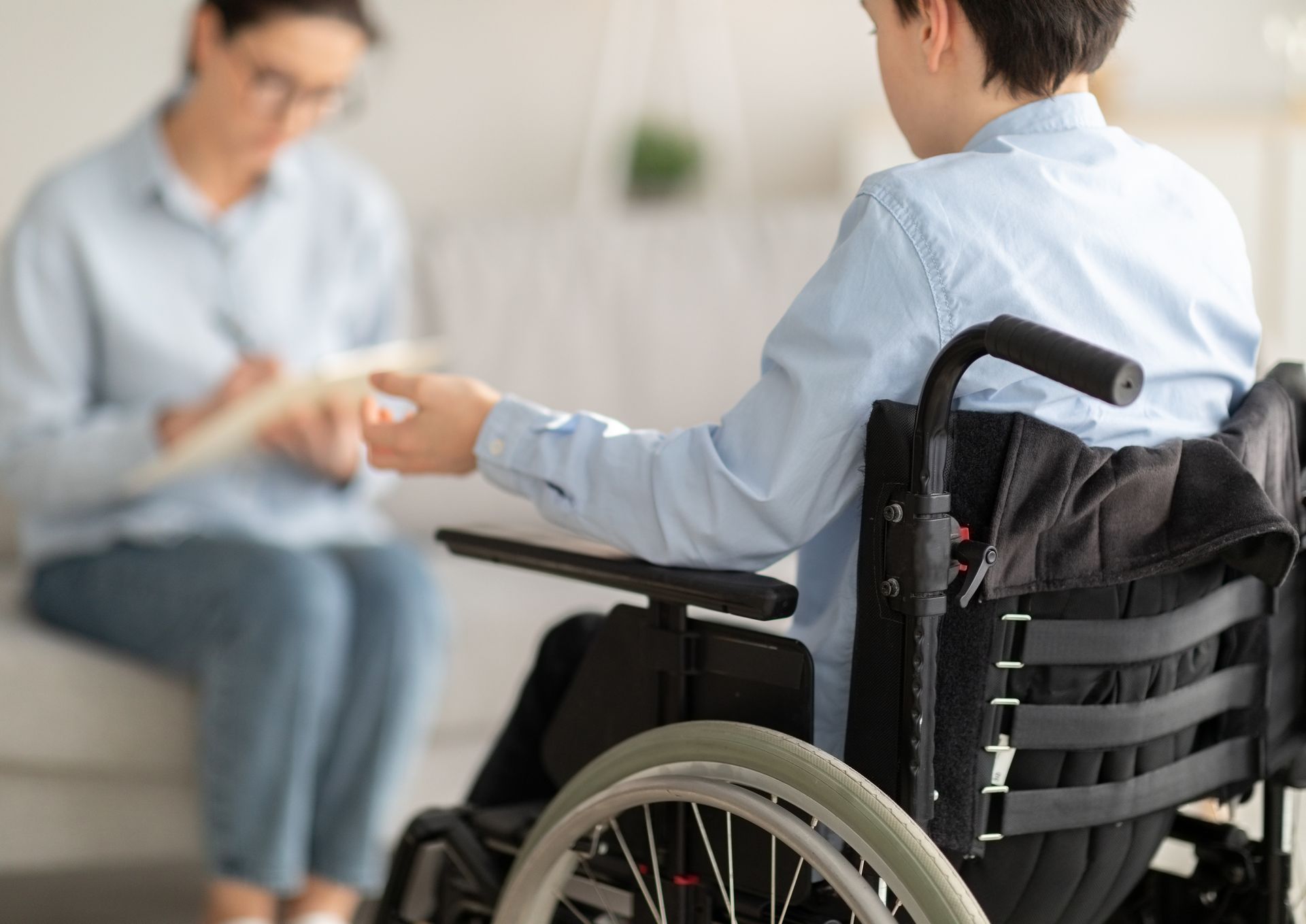 A therapist sits on a couch taking notes while listening to a person sitting in a wheelchair during a consultation.