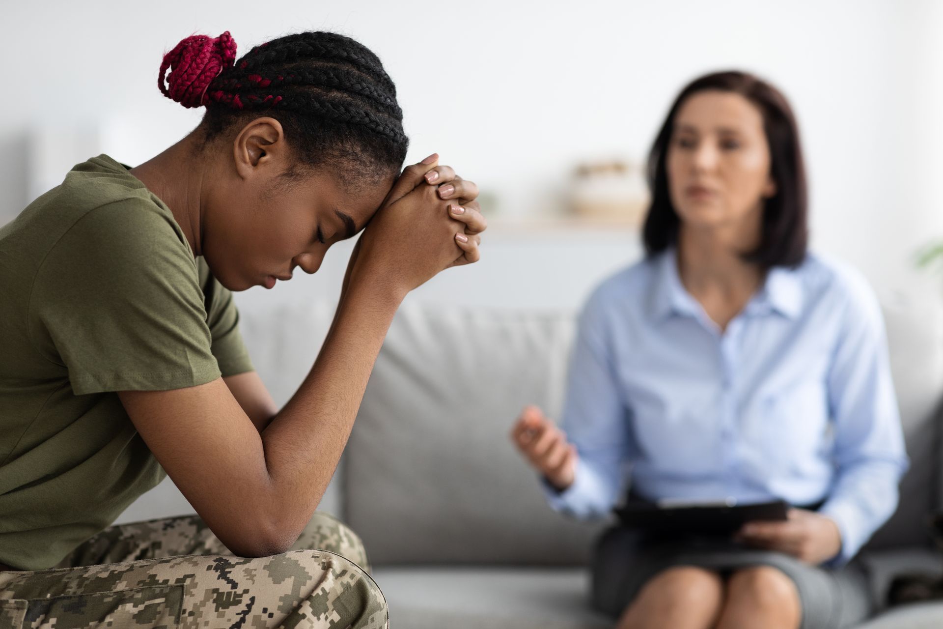 A soldier in uniform sits on a sofa, head in hands, during a counseling session with a professional taking notes.