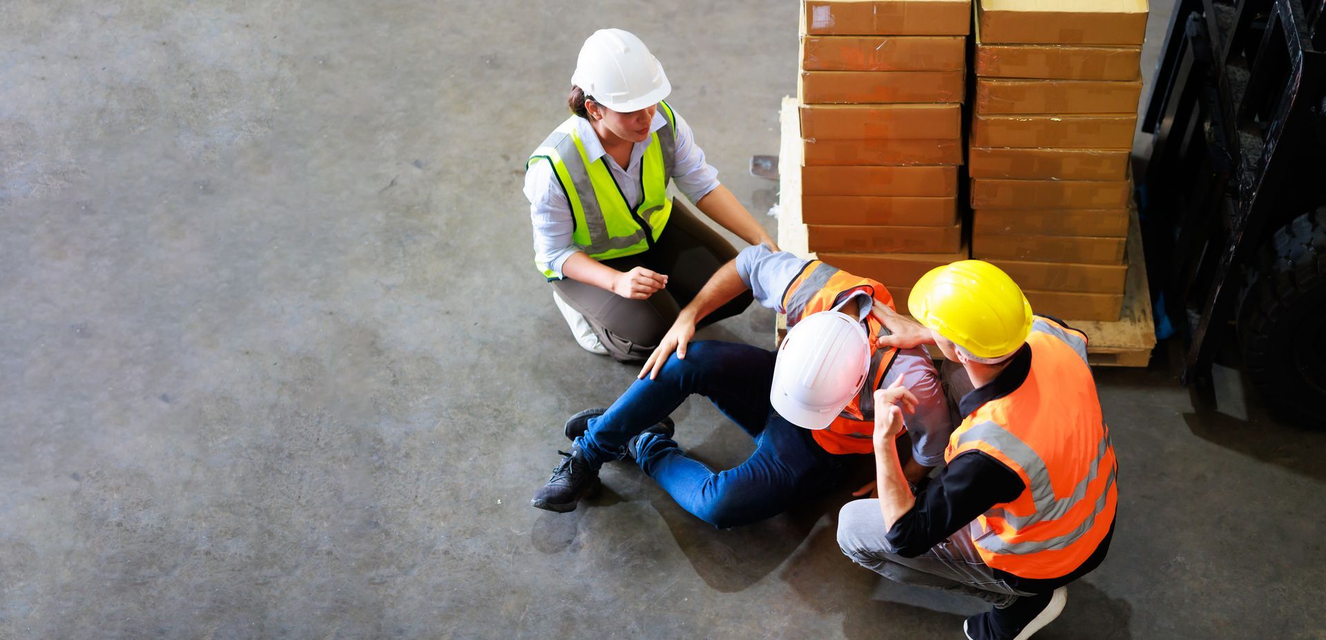 Two workers in safety gear assist a colleague sitting on the warehouse floor near a stack of cardboard boxes.