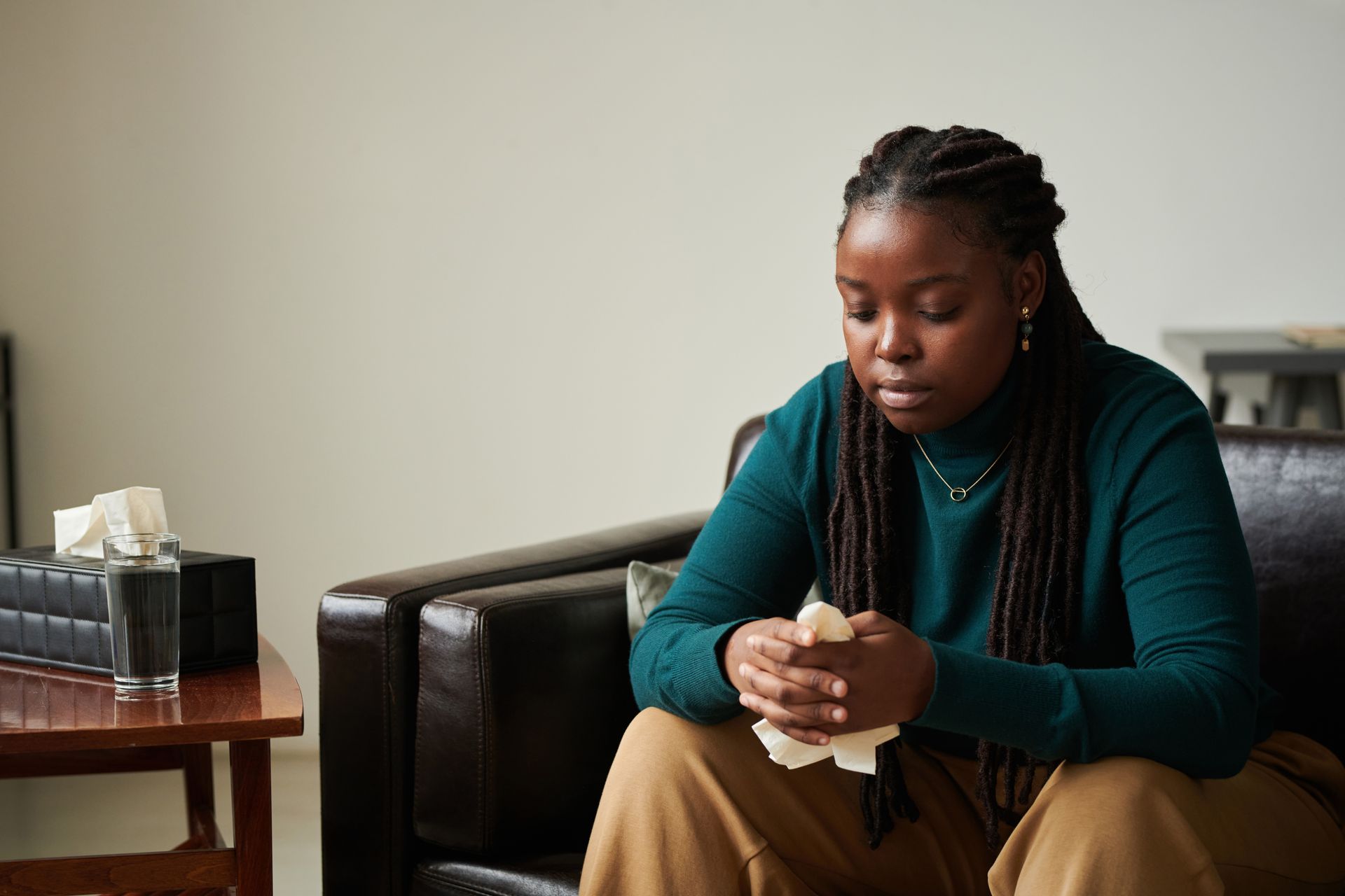 A person in a dark shirt sits on a gray couch, hands clasped near their face, looking thoughtful in a bright room.