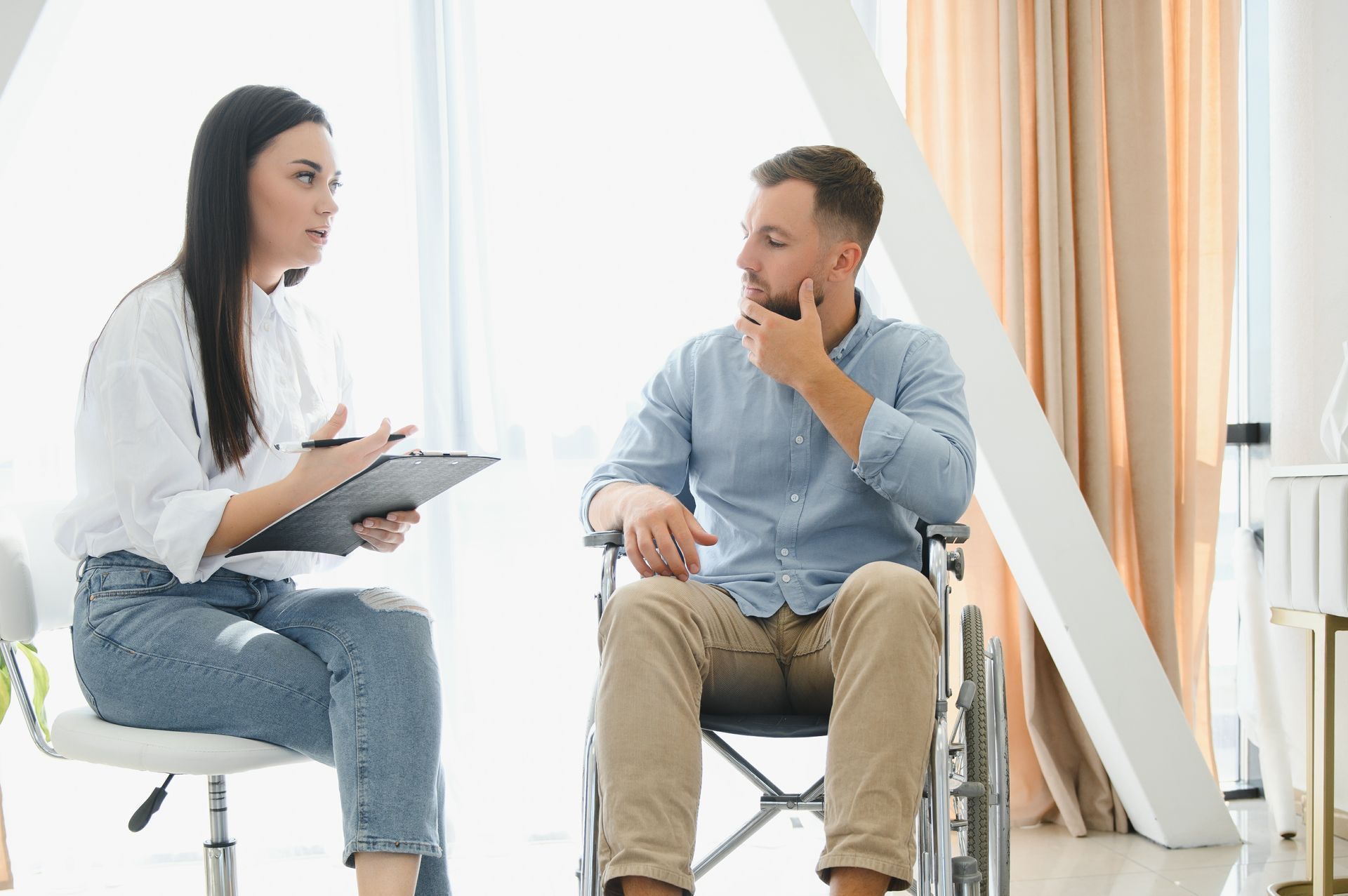 A professional takes notes while talking to a person seated in a wheelchair in a bright, modern office.