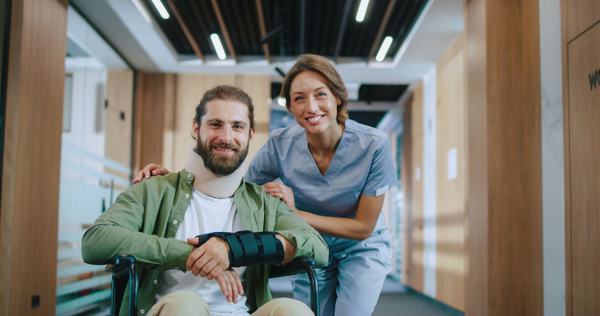 A person in a wheelchair with a neck brace and wrist splint smiles next to a healthcare professional in a hallway.