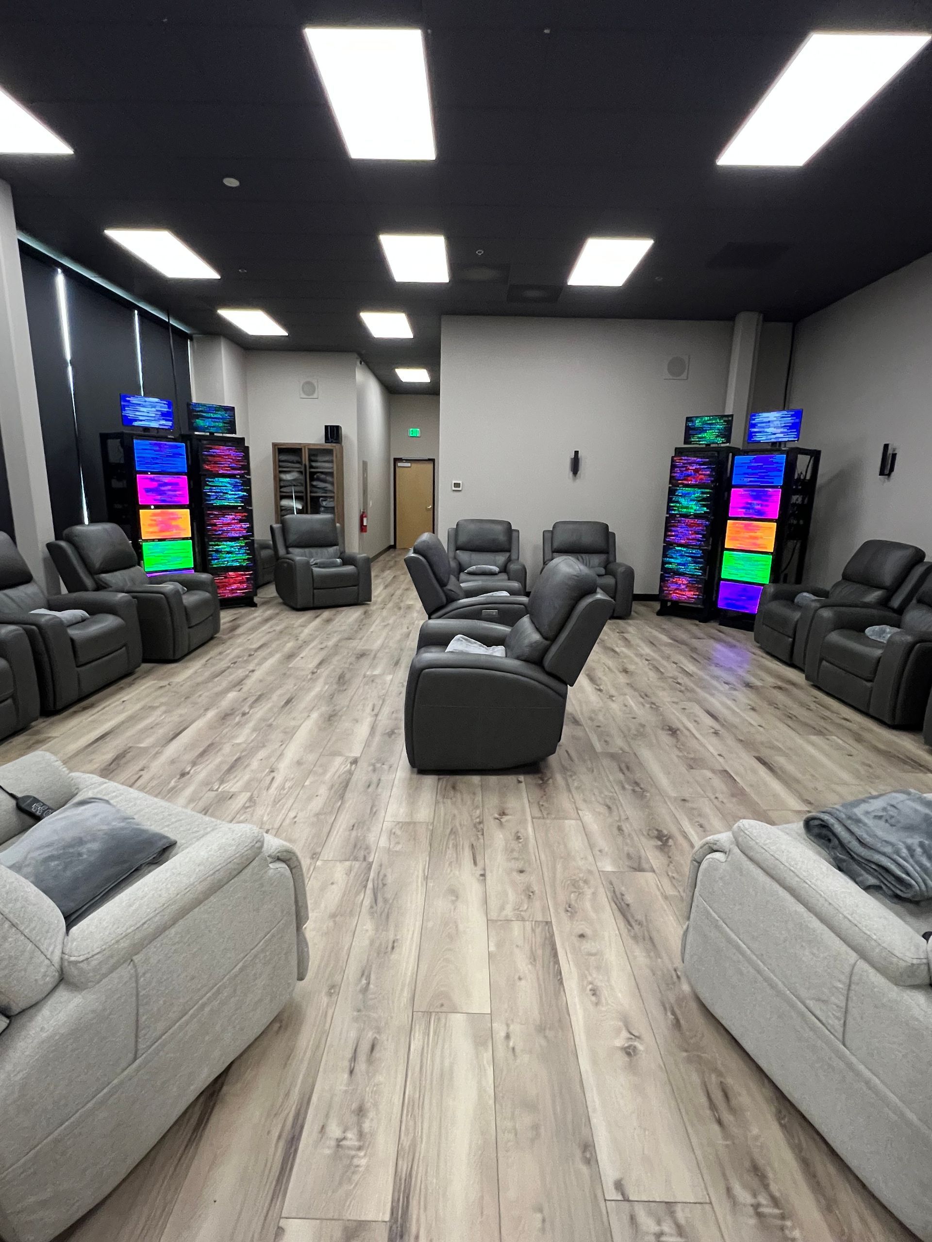 Room with gray recliners and couches. Colorful lit gaming towers stand against the walls; light-colored wood floor.