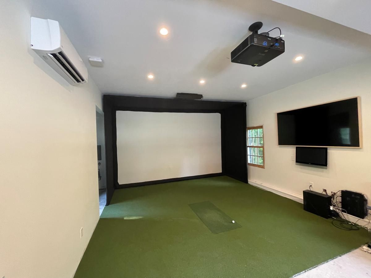 Golf simulator room with a screen, projector, TV, green turf, and air conditioner.