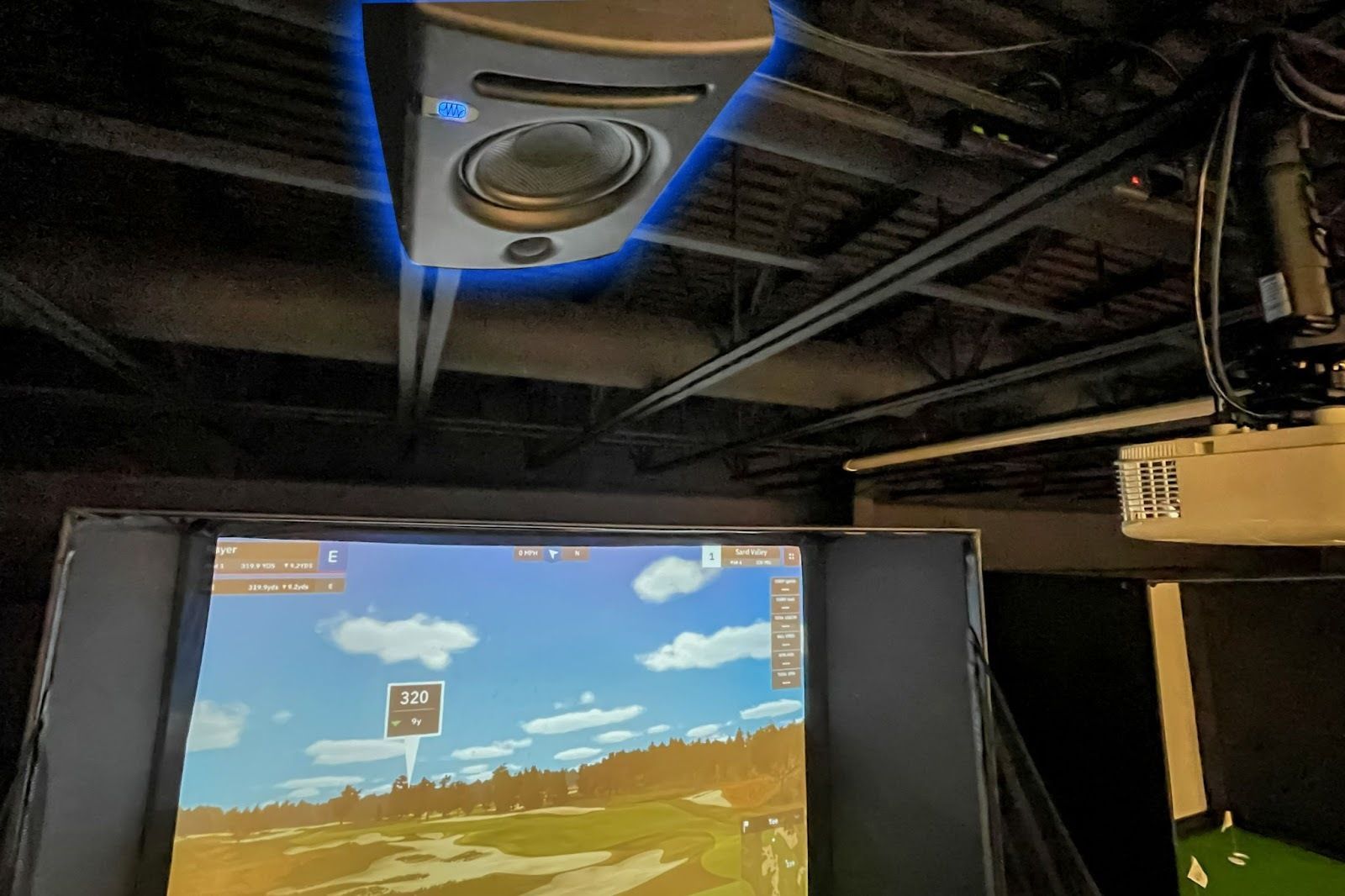 Golf simulator setup: Ceiling speaker, screen displaying golf course, projector, and metal support beams.