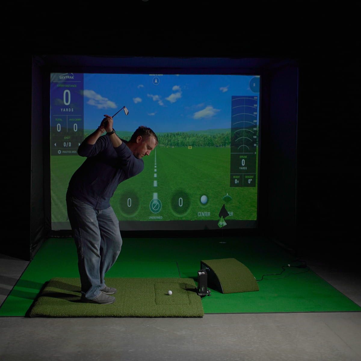 Golf simulator setup with a screen displaying a golf course, projector, and golf bag.