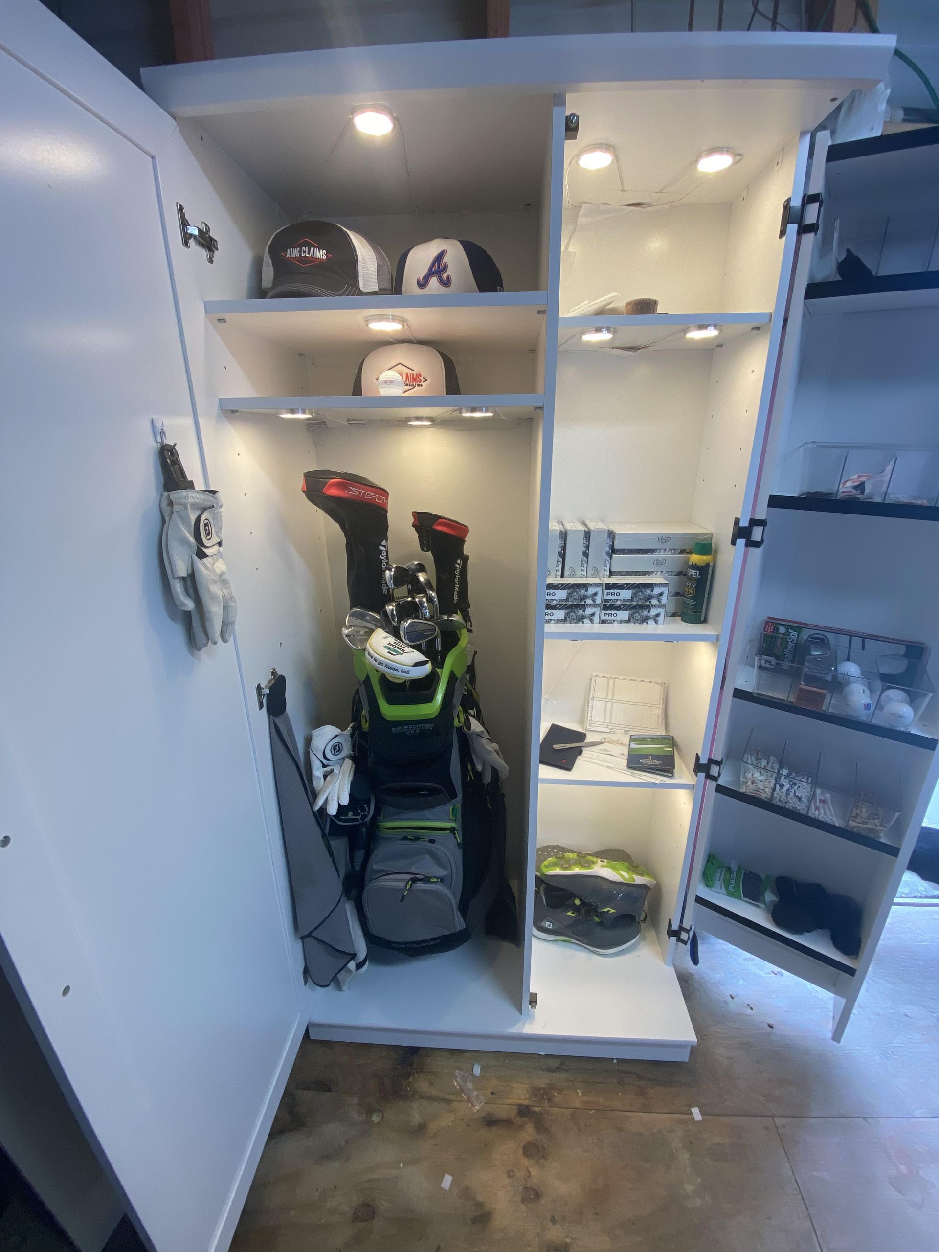 White golf storage cabinet with clubs, hats, shoes, and accessories, illuminated by overhead lights.