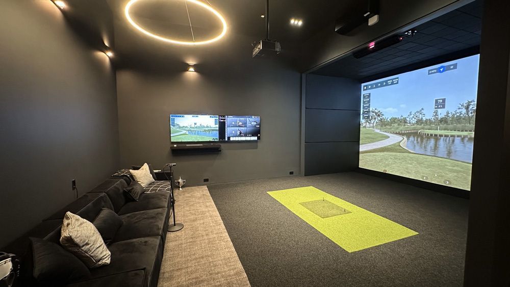 Golf simulator room with large screen, dark walls, sofa, and green turf.