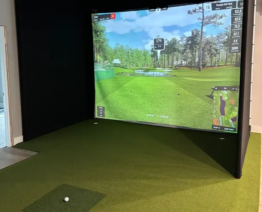 Golf simulator setup showing a large screen projecting a golf course scene, on green turf, with a golf ball on the ground.