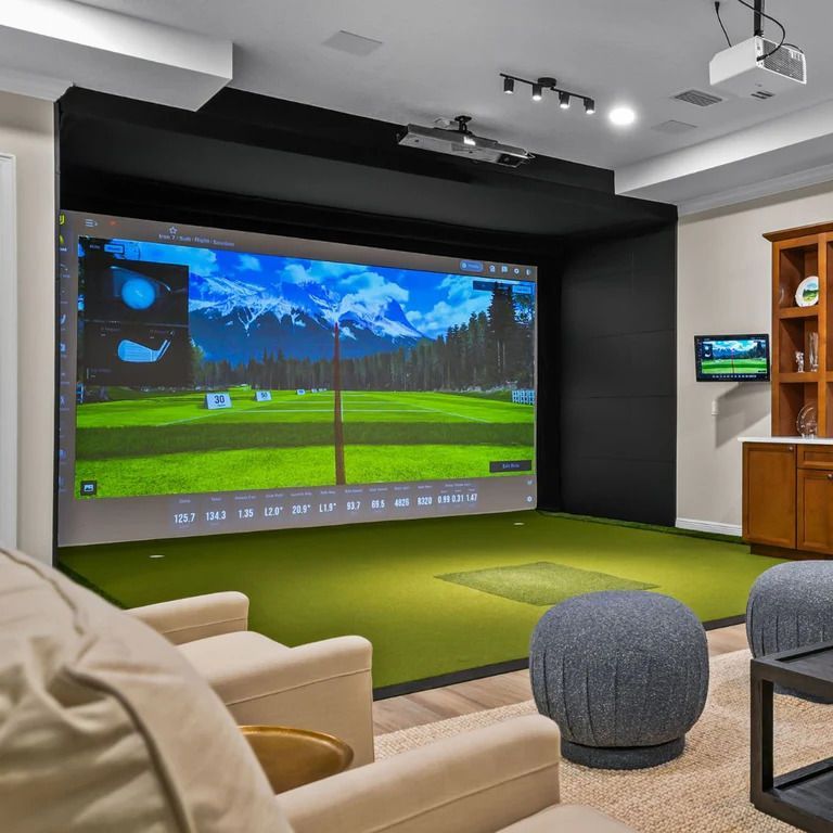 Golf simulator room with screen displaying golf course, green turf, seating.