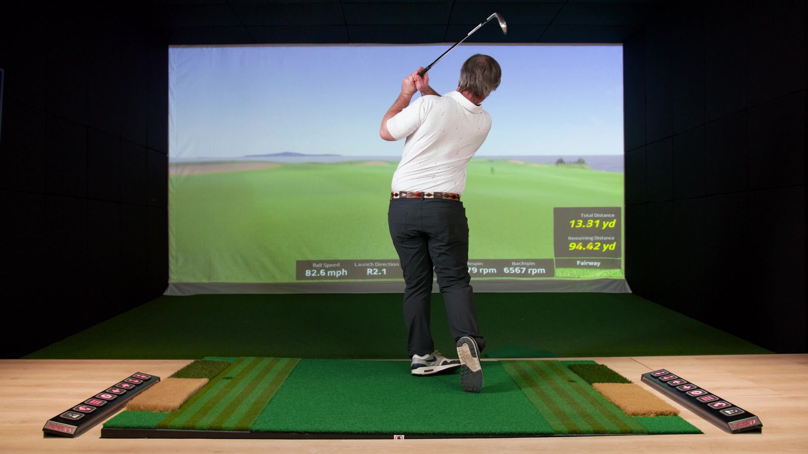 Man swinging a golf club in a golf simulator. He's on a green mat with a screen showing a course.