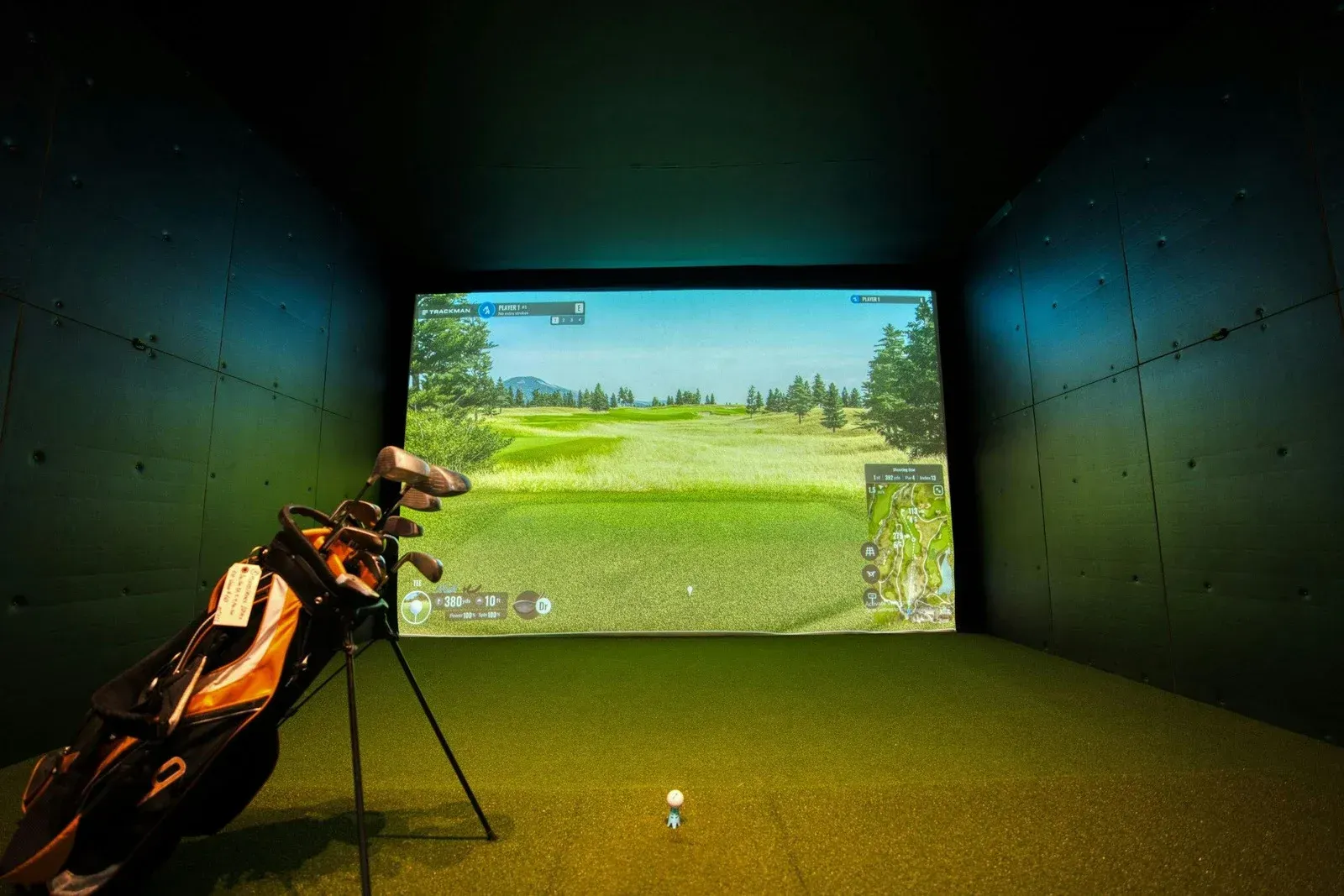 Golf simulator with golf bag, ball, and screen displaying a golf course. Green and black interior.