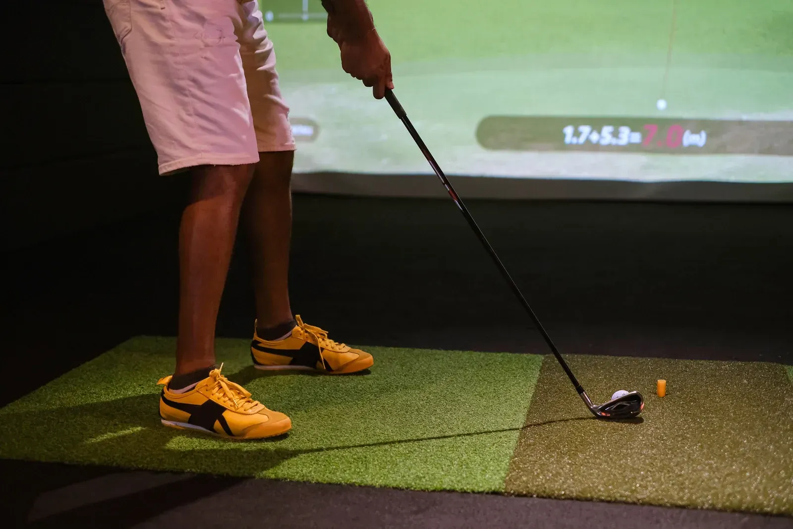 Golfer swinging a club in an indoor golf simulator. They wear shorts and yellow shoes, hitting a ball.