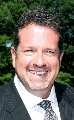 Man in suit smiles at the camera with green foliage in the background.