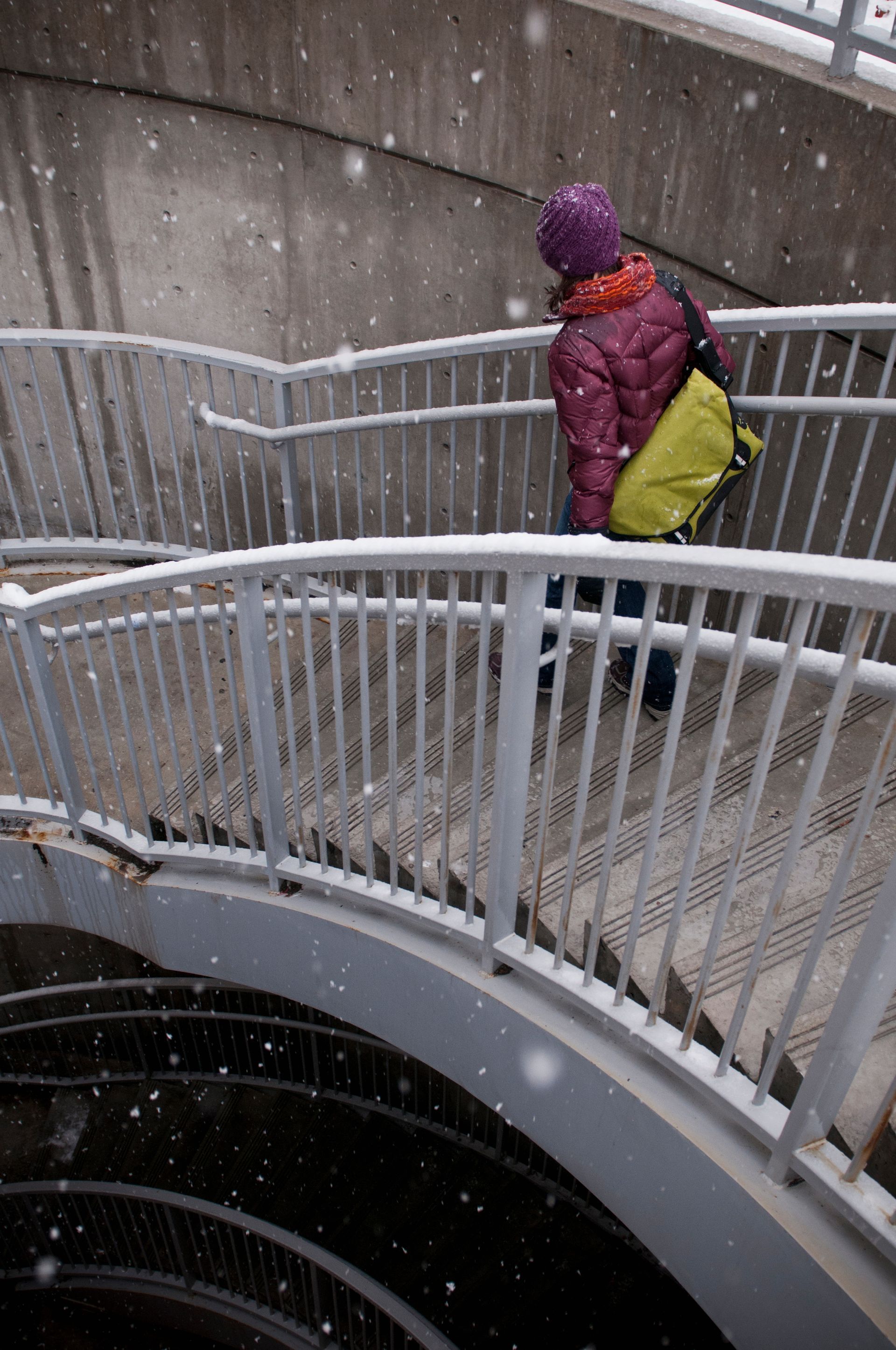 Person in purple hat descends snowy, curved staircase. Yellow bag, orange scarf, gray railings, concrete setting.
