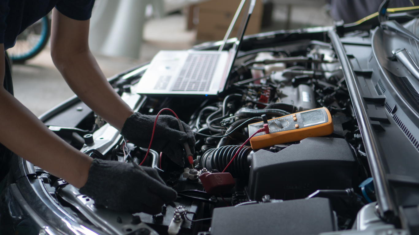 Mechanic working on a car engine with a laptop and multimeter in a garage.