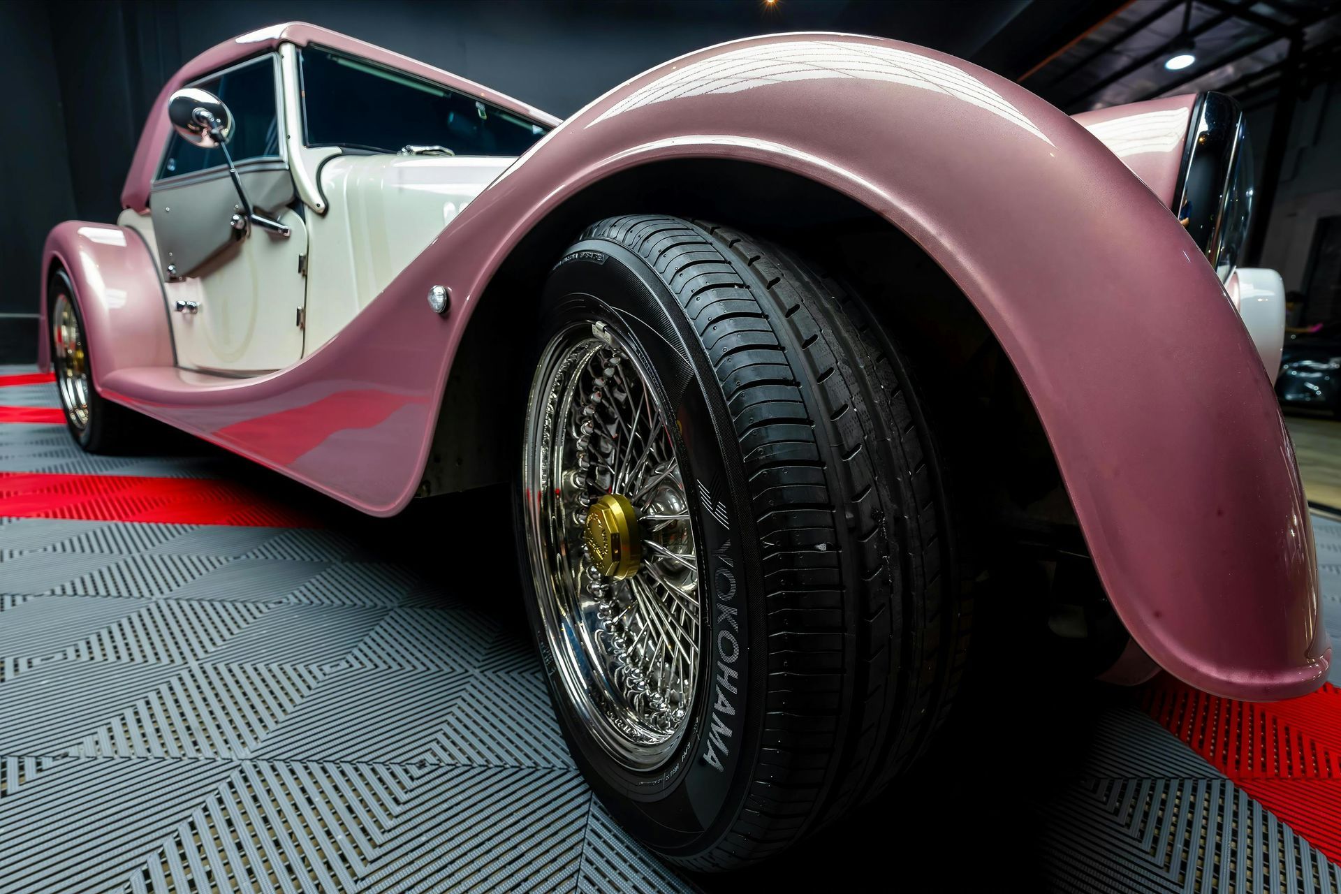 Pink and white vintage car with chrome wheels parked on a checkered garage floor.