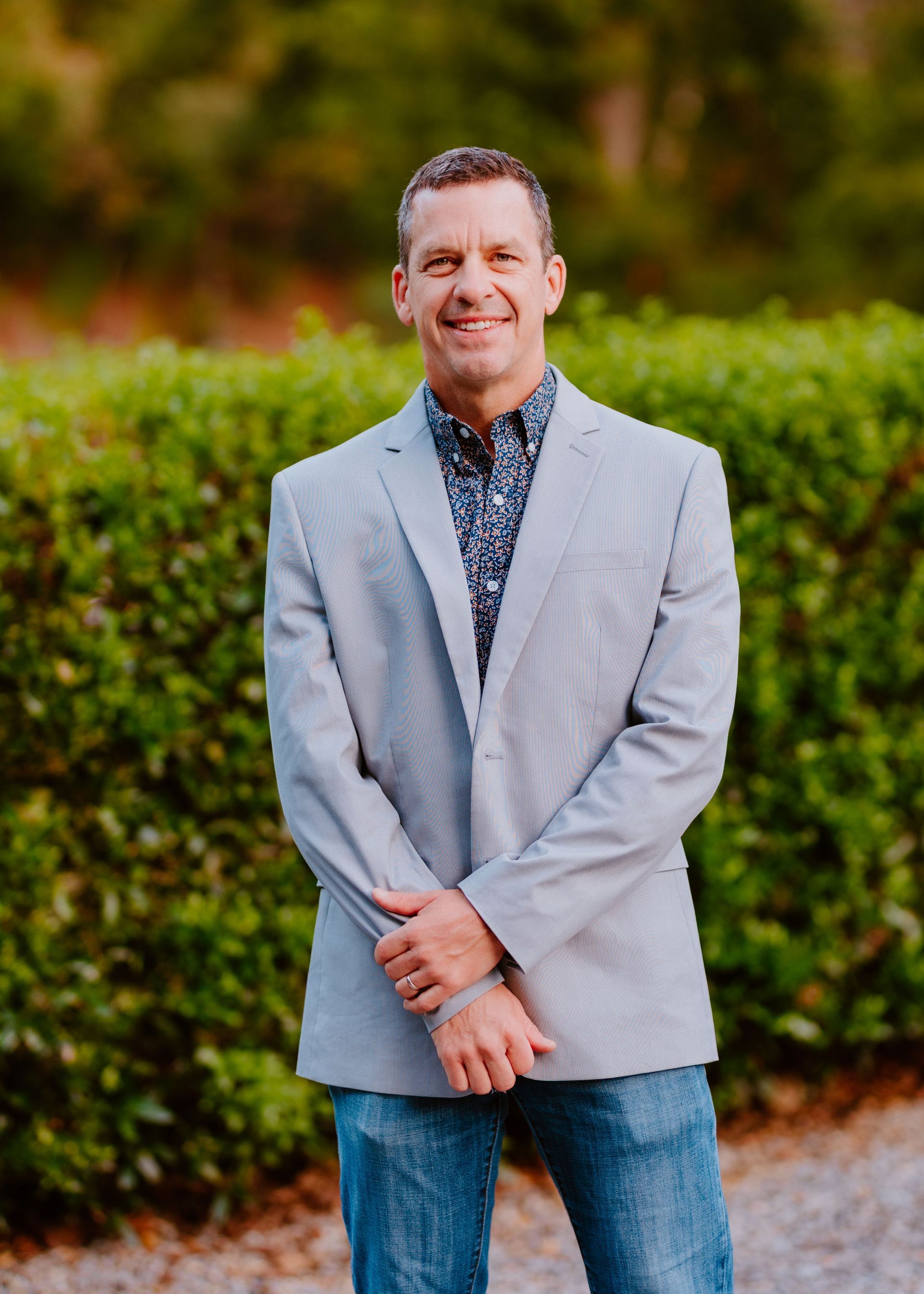 Man in a gray blazer and jeans smiles in front of a green hedge.