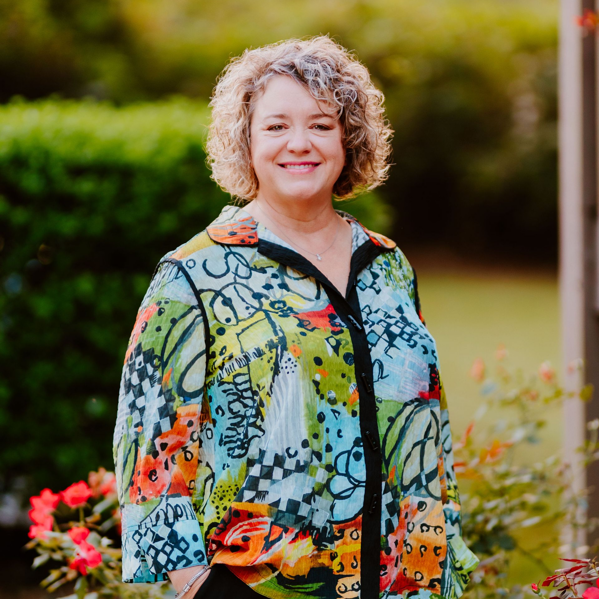 Woman with curly hair smiles outdoors, wearing a colorful patterned top.