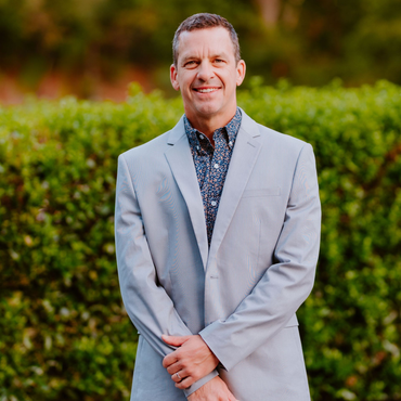 Man in a light gray suit and patterned shirt smiling in front of a green hedge.