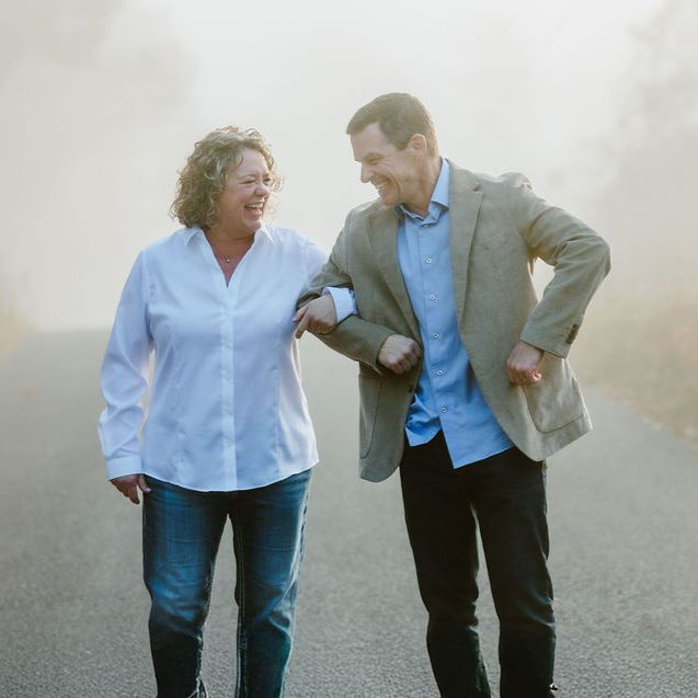 Two people walking, arm in arm, laughing, on a foggy road. One wears a blazer, the other, a white shirt and jeans.