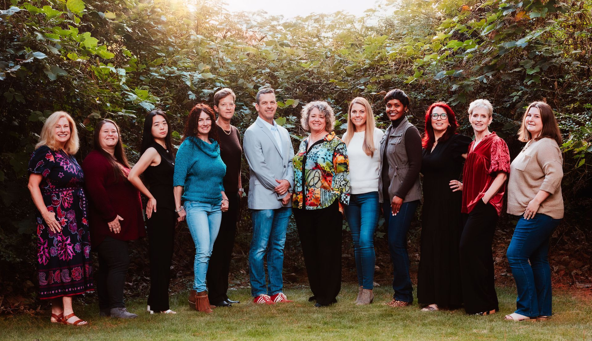 Group of people standing outdoors, smiling. Green foliage background, warm sunlight.