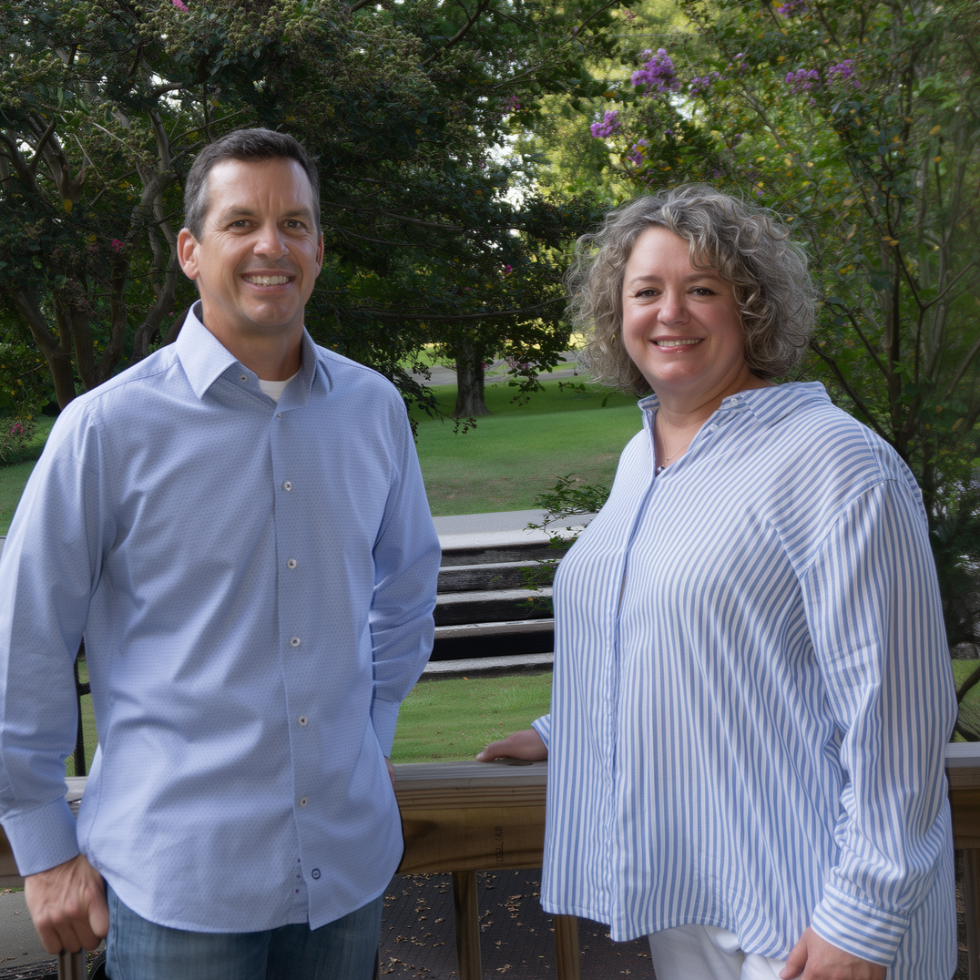 A man and woman stand on a deck smiling, trees and grass in the background.