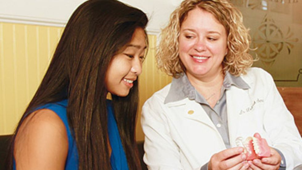 A woman is sitting next to a dentist holding a model of teeth.