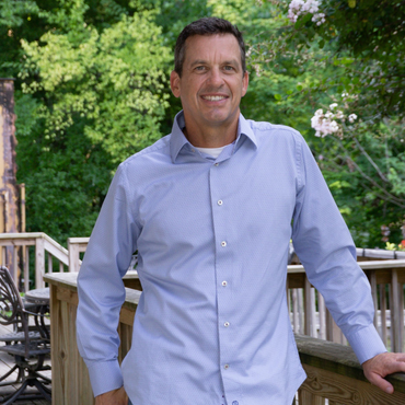 Man in blue shirt smiles, leaning on a deck railing, trees in background.