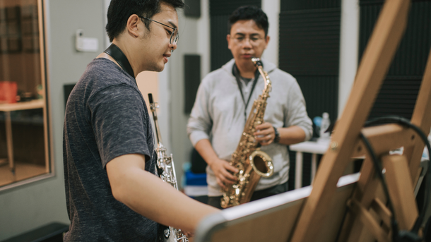 Two men playing saxophones in a music studio. One is reading sheet music, the other looks on.