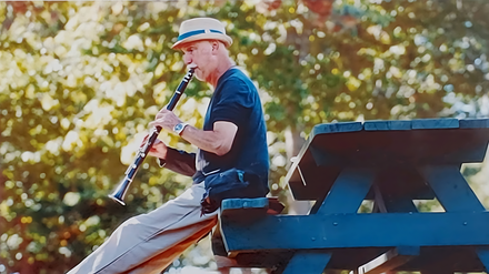 Man in a hat playing a clarinet while seated on a picnic table, outdoors.